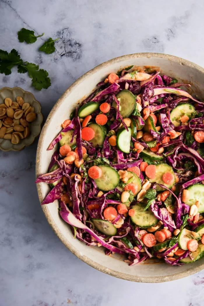 A vibrant Crunchy Red Cabbage Salad in a bowl features sliced cucumbers, carrots, red cabbage, and peanuts, all mixed with fresh cilantro. To the side, there's a small dish containing more peanuts. The light marbled surface background enhances the colorful ingredients.