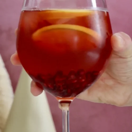 A hand holds a stemmed glass filled with a deep red Festive Aperol Spritz, adorned with floating orange slices and visible pomegranate seeds. The backdrop is a soft pink, with blurred shapes hinting at decorative objects or perhaps a softly blurred Christmas tree shape below.