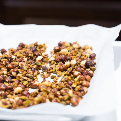 A close-up of a baking sheet lined with parchment paper, filled with spicy candied nuts, including hazelnuts and pistachios. The nuts are golden brown, indicating they are well-roasted. The background is softly blurred, emphasizing the texture and fiery glaze of the flavorful nuts.