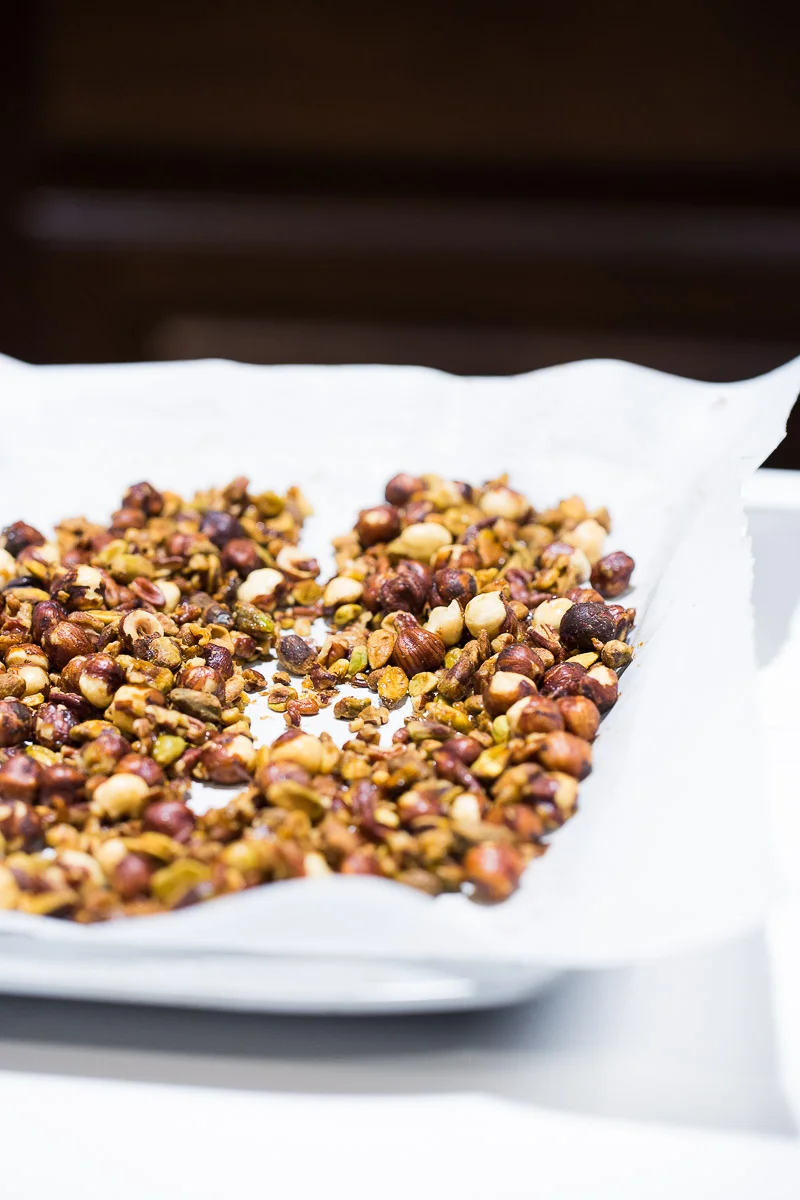 A close-up of a baking sheet lined with parchment paper, filled with spicy candied nuts, including hazelnuts and pistachios. The nuts are golden brown, indicating they are well-roasted. The background is softly blurred, emphasizing the texture and fiery glaze of the flavorful nuts.