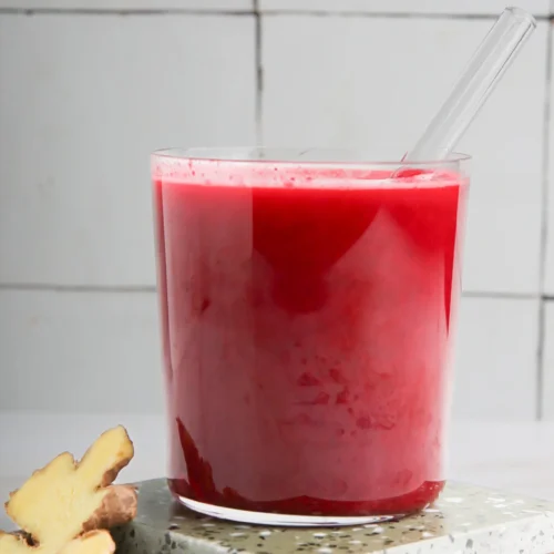 A vibrant beet juice in a clear glass on stacked terrazzo coasters. A glass straw is inserted into the juice. Slices of fresh ginger are placed beside the coasters. The background is a white tiled wall, creating a clean and minimalistic look.