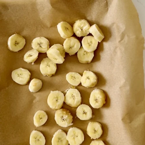Sliced banana pieces, ready for freezing bananas, are spread unevenly on crumpled brown parchment paper. The paper rests on a light, textured surface. The circular slices reveal their soft, creamy interior with tiny seed specks at the center.