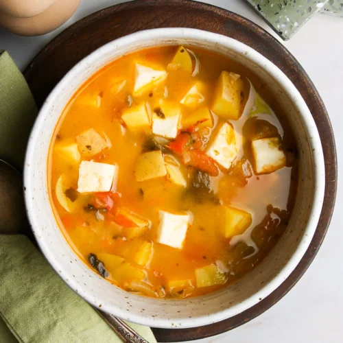A bowl of vibrant vegetable soup with cubes of tofu in a creamy broth, reminiscent of Caldo de queso, is garnished with chopped herbs. Served in a white ceramic bowl on a wooden plate, a green napkin and vintage spoon lie nearby. A pepper shaker and green cup complete the cozy setting.