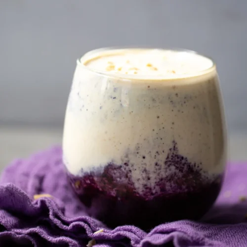Un vaso transparente lleno de un batido en capas reposa sobre un paño morado doblado. La capa inferior es de color morado oscuro, probablemente de frutos rojos, y la superior es de color beige cremoso, con una pizca de nueces por encima. El fondo gris está suavemente difuminado, resaltando el batido.