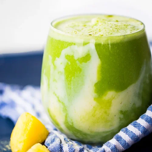 A clear glass brimming with a vibrant green smoothie, infused with matcha and swirled with creamy yellow, graces a blue surface adorned by a striped cloth. In the foreground sit three chunks of yellow mango. The softly blurred background enhances its fresh and inviting appeal.