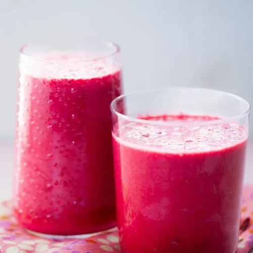Two clear glasses filled with vibrant Pink Power Smoothies sit on a colorful floral napkin. The nutrient-packed beverages boast a frothy texture on top, with small bubbles visible. The background is softly blurred, accentuating the bright and inviting appearance of the drinks.