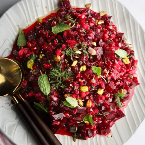 A white plate holds a vibrant beet salad topped with pomegranate seeds, mint leaves, pistachios, and dill sprigs. Two black-handled serving spoons rest beside the plate. A pink and white striped cloth is partially visible in the bottom left corner on a light marble surface.