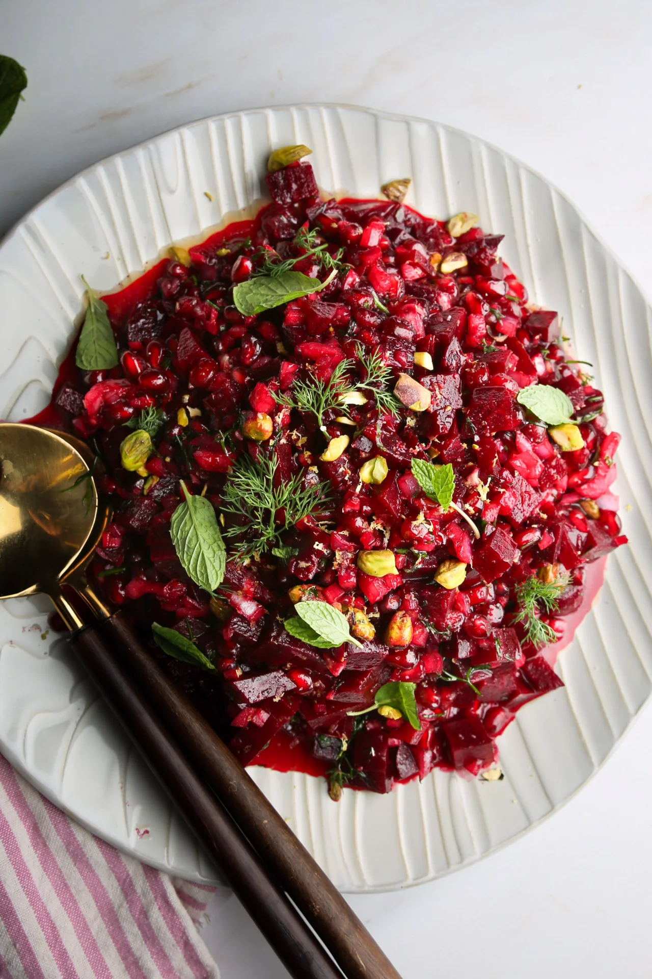 A white plate holds a vibrant beet salad topped with pomegranate seeds, mint leaves, pistachios, and dill sprigs. Two black-handled serving spoons rest beside the plate. A pink and white striped cloth is partially visible in the bottom left corner on a light marble surface.