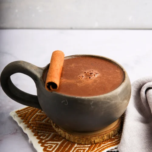 A rustic ceramic mug filled with frothy Mexican hot chocolate sits on a patterned, woven coaster. A cinnamon stick rests on the mug's rim. A light gray napkin is placed beside it on a marble surface, while a smooth, neutral backdrop frames the cozy scene.