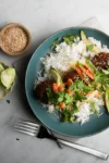 A blue plate features a twist on banh mi with white rice topped with savory meatballs, avocado slices, carrot strips, cilantro, and sesame seeds. Two lime halves add zest nearby. A small wooden bowl of sesame seeds, alongside a fork and knife, complete the scene on a marble surface.