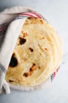 A stack of freshly made homemade flour tortillas wrapped partially in a white cloth with red and blue stripes. The flatbreads have a light golden-brown color with darker brown spots, indicating they were cooked on a hot surface, and are placed on a light marble countertop.
