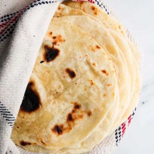 A stack of freshly made homemade flour tortillas wrapped partially in a white cloth with red and blue stripes. The flatbreads have a light golden-brown color with darker brown spots, indicating they were cooked on a hot surface, and are placed on a light marble countertop.