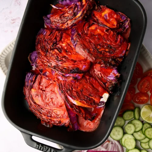 Grilled cabbage slices in a black baking dish, charred with a reddish glaze reminiscent of sarandeada sauce. Beside the dish, a white platter holds sliced cucumbers, red onions, tomatoes, and lime wedges, all arranged beautifully on a marbled surface.