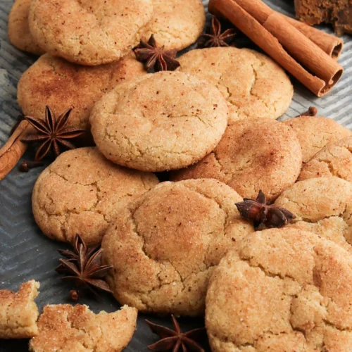 A batch of golden-brown snickerdoodle cookies is arranged on a textured gray surface. The cookies are sprinkled with cinnamon sugar. Cinnamon sticks and star anise are scattered around, along with a broken cookie piece in the foreground. Dark chocolate rests in the top corner.