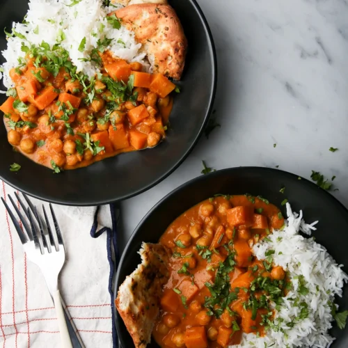 Two black bowls filled with a vibrant chickpea and sweet potato curry garnished with fresh cilantro, served with white rice and a piece of naan. The bowls are on a white marble surface alongside a folded cloth napkin with a red check pattern and two forks.