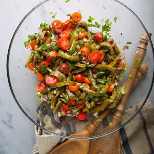 A clear glass bowl brimming with an easy recipe of green beans, cherry tomatoes, and purple onions, garnished with cilantro. Resting on a beige cloth with blue stripes, the bowl is accompanied by two wooden serving utensils on a marble countertop.