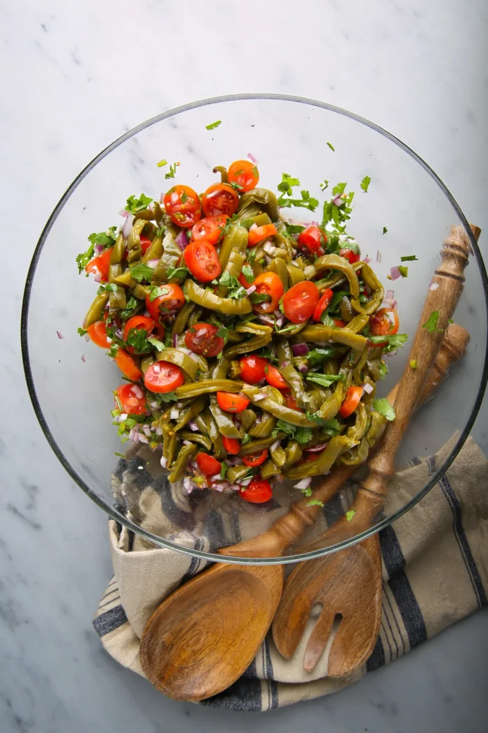 A clear glass bowl brimming with an easy recipe of green beans, cherry tomatoes, and purple onions, garnished with cilantro. Resting on a beige cloth with blue stripes, the bowl is accompanied by two wooden serving utensils on a marble countertop.