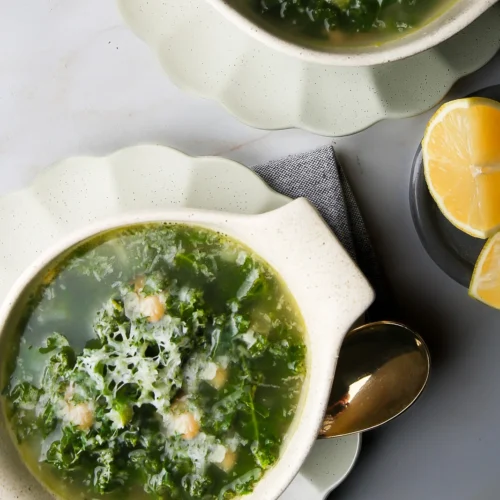 Two bowls of kale and white bean soup topped with grated cheese, placed on scalloped white plates. A golden spoon and a linen napkin are beside one bowl. Lemon wedges on a small plate are on the side. The background is a light gray surface.