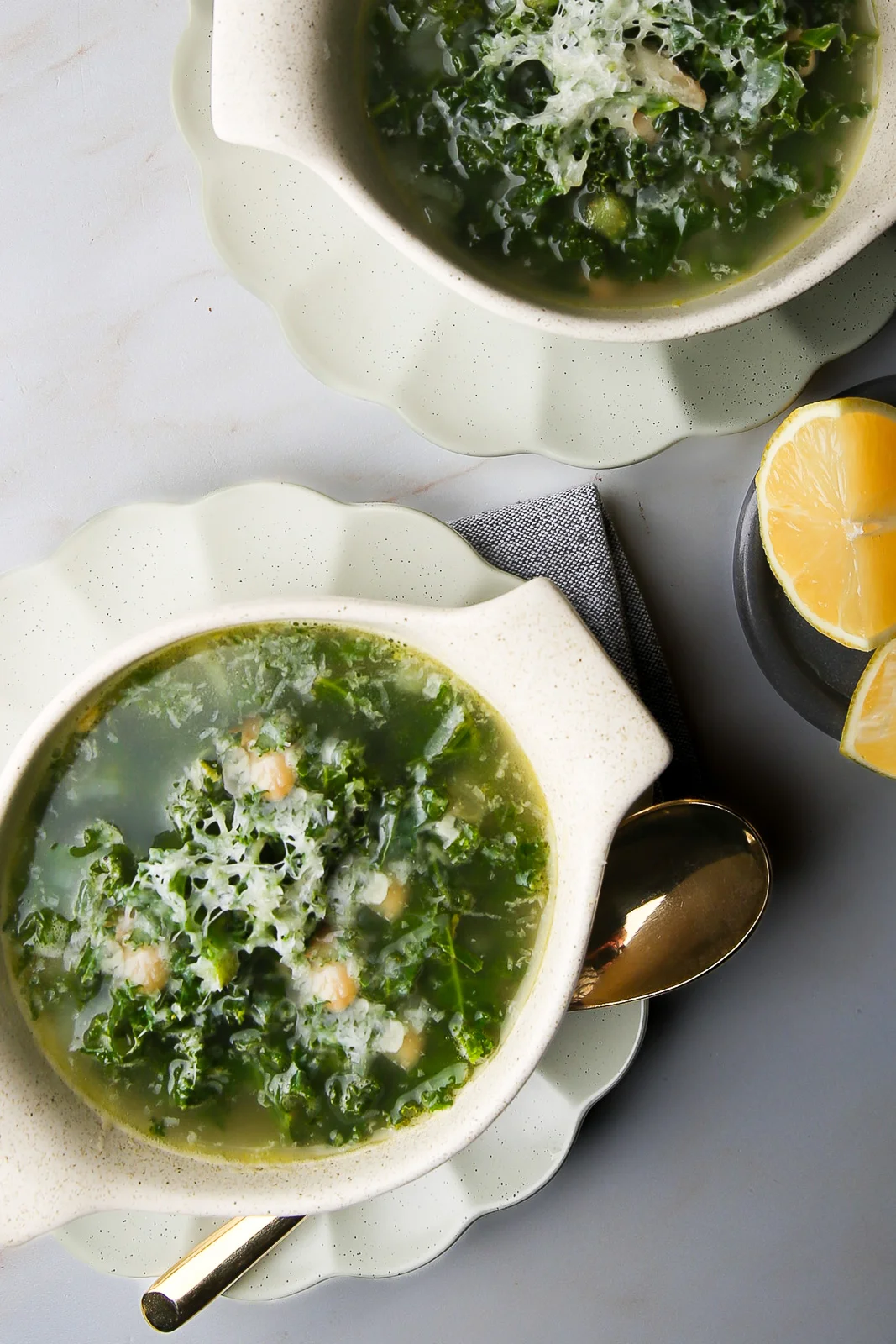 Two bowls of soup sit on a light gray surface. Each bowl contains a green broth infused with garlic and leafy kale, garnished with grated cheese and small pieces of shrimp. A gold spoon rests beside one bowl. Two lemon wedges are placed on a small dark plate nearby.