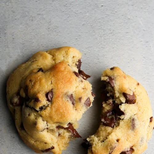 Two Levain Bakery-Style chocolate chip cookies rest on a gray surface, one broken in half. These thick, eggless delights boast gooey chocolate chunks and a golden-brown hue, suggesting they are freshly baked and irresistibly soft.