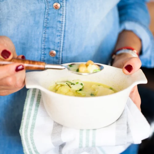 A person in a blue denim shirt holds a white ceramic bowl filled with a creamy soup. The bowl rests on a striped cloth. The person uses a spoon with a wooden handle to scoop the soup, adorned with herbs, possibly parsley. Their nails are painted dark red.