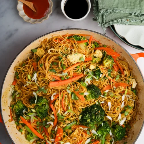 A large pan filled with stir-fried noodles, broccoli, carrots, and tofu. The noodles are mixed with sliced green onions and sauce. To the side, there's a small bowl of soy sauce, a dish of chili sauce with a spoon, and a stack of green napkins with wooden chopsticks on a marble surface.