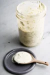 A glass jar filled with creamy ranch dressing is open, with some dressing visible on the inner sides. A small ceramic spoon rests on a round, dark gray plate, holding a dollop of the dressing. The dressing appears smooth and is speckled with herbs. The background is a light surface.