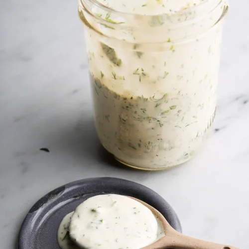 A glass jar filled with creamy ranch dressing is open, with some dressing visible on the inner sides. A small ceramic spoon rests on a round, dark gray plate, holding a dollop of the dressing. The dressing appears smooth and is speckled with herbs. The background is a light surface.