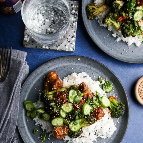 A gray plate with white rice topped with glazed chicken, broccoli, sliced cucumbers, and sprinkled sesame seeds. Next to it is another similar plate. A glass of sparkling water sits on a speckled coaster. A can sits nearby next to a folded gray cloth with utensils on it.