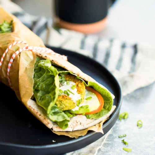 A wrapped falafel sandwich on a black plate features crispy chickpea falafel balls, lettuce, cucumber slices, and tomato encased in pita bread. Partially covered with brown paper tied with string, it sits against a backdrop of scattered cilantro garnishes and a striped cloth.