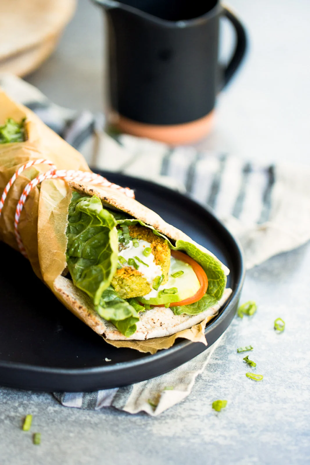 A wrapped falafel sandwich on a black plate features crispy chickpea falafel balls, lettuce, cucumber slices, and tomato encased in pita bread. Partially covered with brown paper tied with string, it sits against a backdrop of scattered cilantro garnishes and a striped cloth.