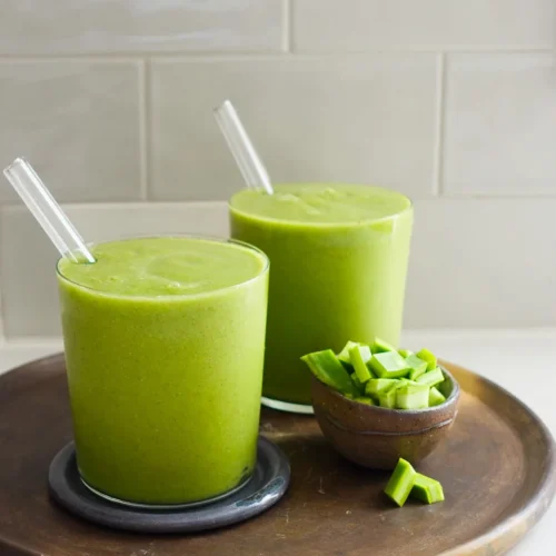 Two glasses of green smoothies with glass straws sit on a round wooden tray, resembling vibrant Mexican green juice. Beside them is a small wooden bowl filled with sliced green vegetables. The background features a tiled wall in light gray, emphasizing the fresh and healthy beverage option.