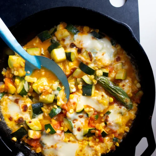 A cast iron skillet holds the savory delight of Calabacitas con queso, featuring Mexican zucchini, corn, and tomatoes mixed with melted cheese. The dish looks saucy and freshly cooked, while a blue serving spoon rests in the skillet on a black square trivet with a circular cutout on top right.