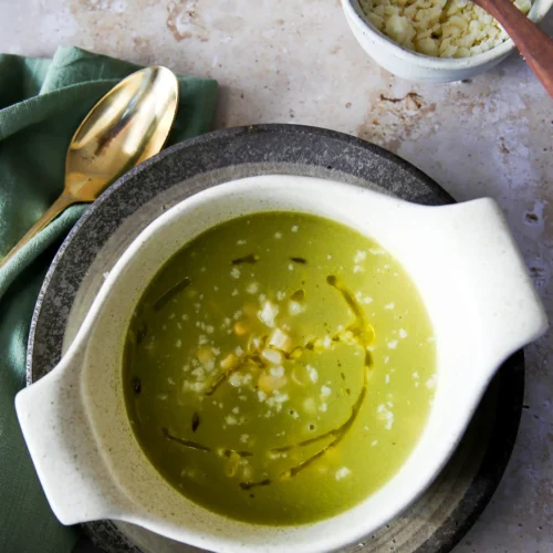 A creamy green Mexican Zucchini Soup garnished with crumbled cheese and a drizzle of olive oil is served in a white bowl. The bowl sits on a dark plate, accompanied by a gold spoon and a green napkin. Nearby, corn rests alongside crumbled cheese in a small bowl with a wooden spoon.