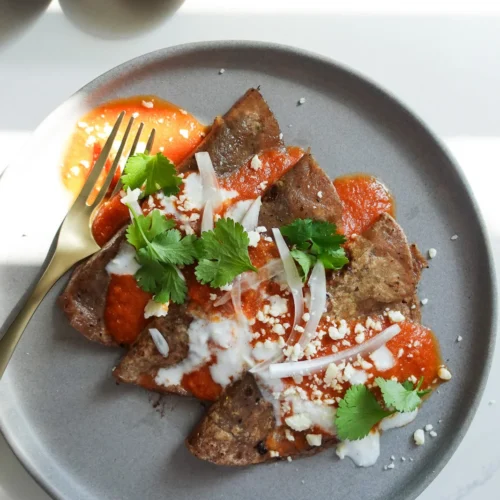 A gray plate with beef liver slices topped with red sauce, crumbled cheese, cilantro, and onion slices. A drizzle of white crema adds contrast. A gold fork rests on the plate. In the background, a small bowl of crumbled cheese is visible on a white surface.