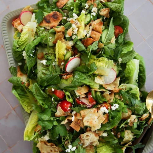 A fresh Fattoush salad on a rectangular tray boasts mixed greens, cherry tomatoes, sliced radishes, crispy pita pieces, herbs, and crumbled feta cheese drizzled with tangy dressing. Tongs are placed nearby. The vibrant mix of colors and textures pops against the tiled background.