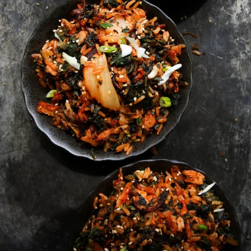 Three bowls of Korean fried rice are set on a dark surface. The dish combines kimchi, seaweed, and green onions, garnished with sesame seeds. Presented on black plates, the vibrant colors of the ingredients create a striking contrast with the muted background.