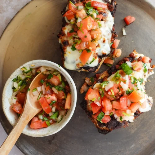A ceramic plate holds two Molletes topped with melted cheese and fresh salsa, including tomatoes, onions, and herbs. Beside the bread, a small bowl contains extra salsa with a wooden spoon, all set on a light stone surface—an authentic touch of Mexican food delight.