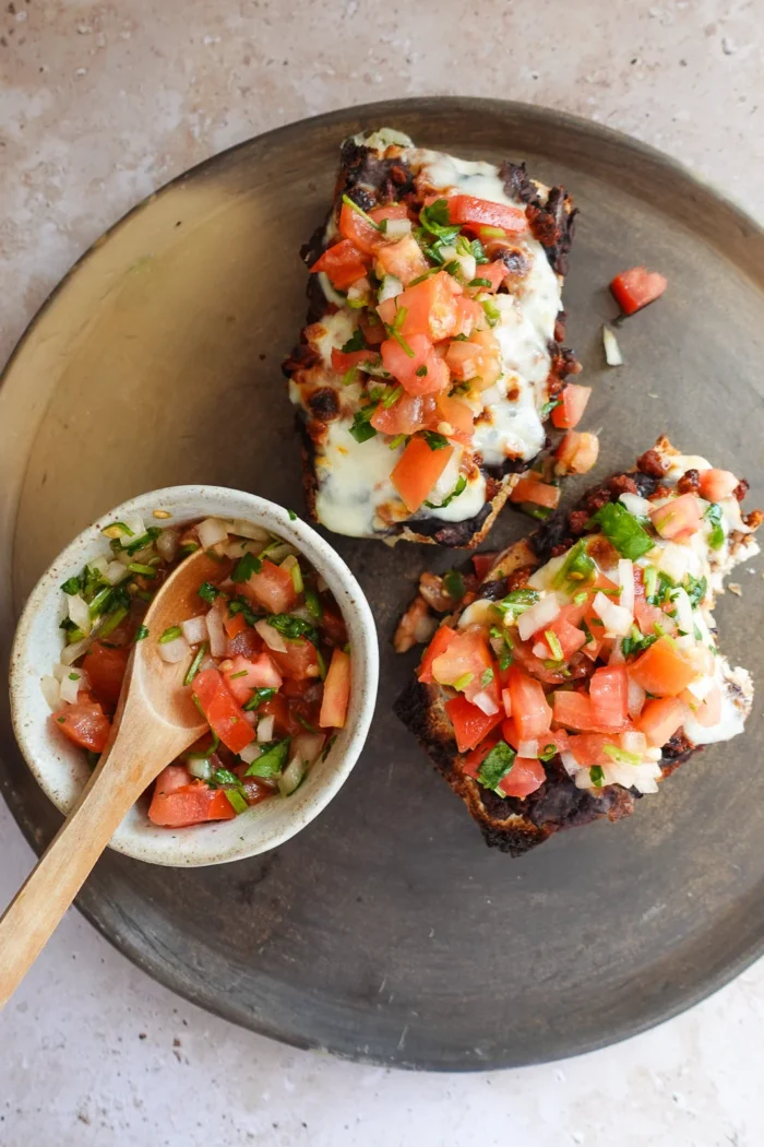 A ceramic plate holds two Molletes topped with melted cheese and fresh salsa, including tomatoes, onions, and herbs. Beside the bread, a small bowl contains extra salsa with a wooden spoon, all set on a light stone surface&mdash;an authentic touch of Mexican food delight.