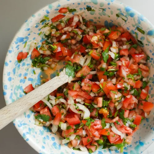 An authentic speckled ceramic bowl brims with vibrant pico de gallo on a light surface. The lively mix includes diced red tomatoes, chopped white onions, green cilantro, and jalapeños. A wooden spoon is partially immersed in the fresh ingredients, offering a rustic touch.