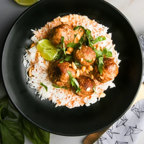A black bowl with rice topped with flavorful meatballs in Thai-style red curry sauce, garnished with chopped green herbs and pine nuts. A lime wedge is on the rice. Lime slices are on the side, along with a patterned napkin and utensils. Fresh basil leaves rest nearby on a marble surface.