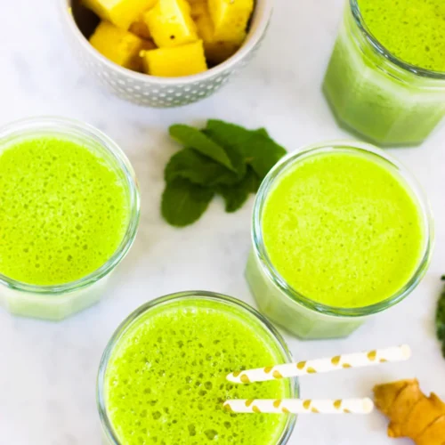Top view of four glasses filled with vibrant green smoothies on a light surface. A small white bowl with pineapple chunks is nearby, along with fresh mint leaves and a piece of turmeric root. Two striped paper straws in one smoothie add a playful touch, making these green smoothies irresistible.