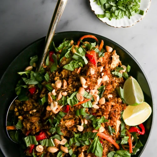 A bowl of fried rice topped with chopped greens, sliced red peppers, and peanuts, garnished with lime wedges on the side. A silver spoon rests in the bowl. In the background, two small plates hold more chopped greens and peanuts. The surface is a gray marble.
