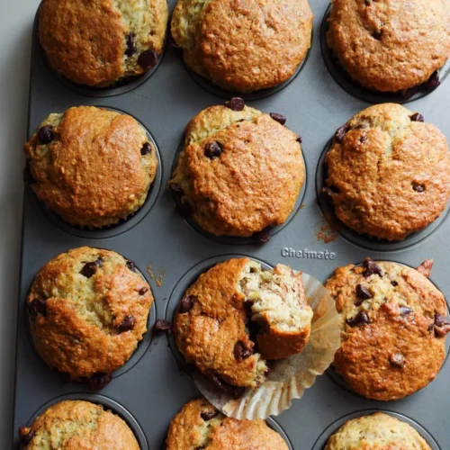 A muffin tray holds twelve golden-brown chocolate chip muffins. One muffin is slightly out of its cup with its wrapper partially peeled back, revealing a soft, moist interior. These eggless delights are set against a light surface, showcasing their tempting homemade texture.