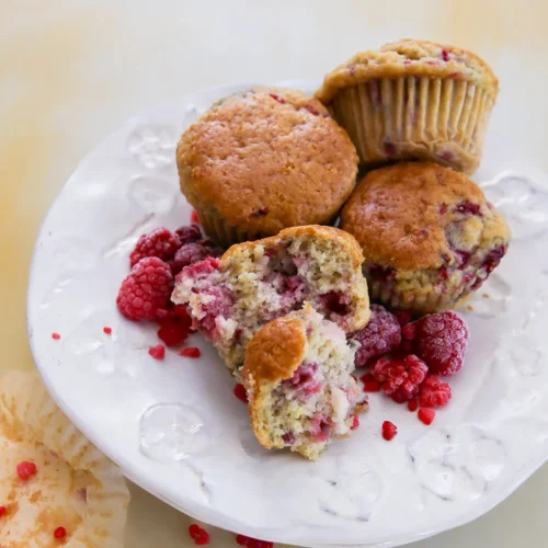 Four raspberry muffins sit on a white plate, one broken open to reveal its moist interior. Fresh and crushed raspberries are scattered around, with a hint of lemon zest adding brightness. An empty muffin wrapper with crumbs lies beside on the light yellow background.