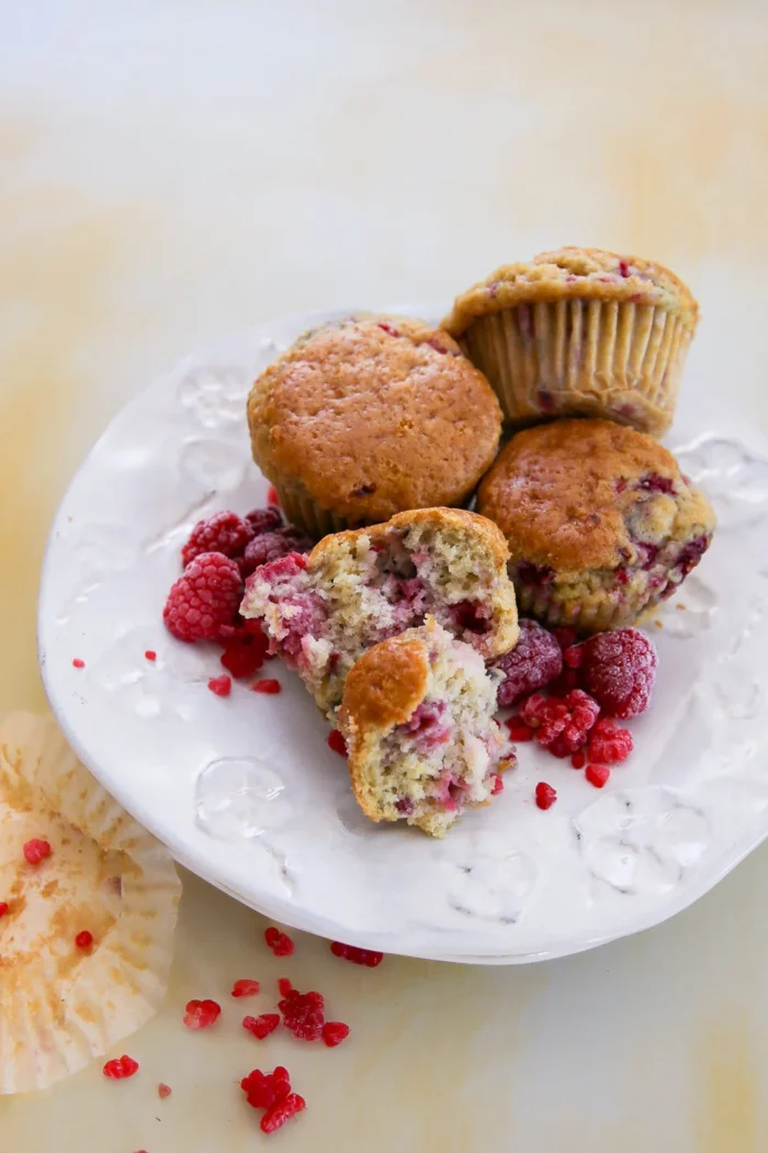 Four raspberry muffins sit on a white plate, one broken open to reveal its moist interior. Fresh and crushed raspberries are scattered around, with a hint of lemon zest adding brightness. An empty muffin wrapper with crumbs lies beside on the light yellow background.