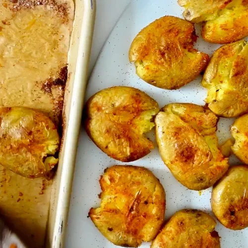 Smashed roasted potatoes with golden, crispy edges are spread on a white plate, evoking classic Patatas Bravas. Seasoned with spices, this Spanish tapas-inspired potato recipe boasts an appetizing, textured appearance next to a baking sheet lined with parchment paper.