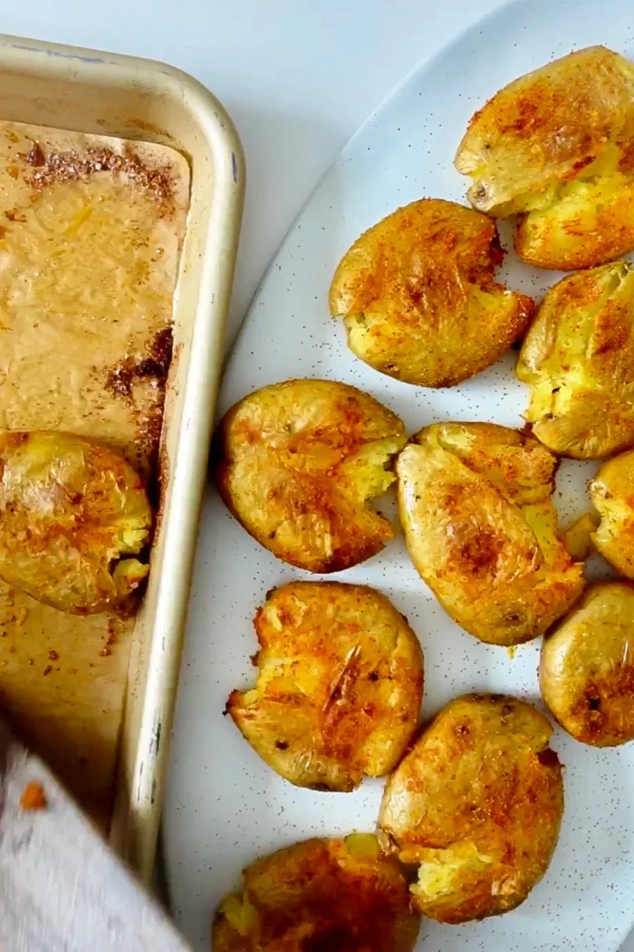 Smashed roasted potatoes with golden, crispy edges are spread on a white plate, evoking classic Patatas Bravas. Seasoned with spices, this Spanish tapas-inspired potato recipe boasts an appetizing, textured appearance next to a baking sheet lined with parchment paper.