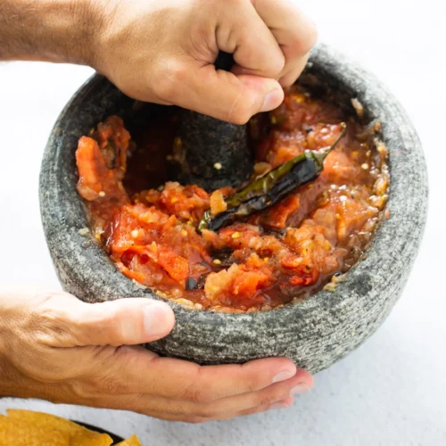 Two hands expertly use a molcajete, the traditional Mexican home cook’s tool, to grind roasted tomatoes, chilies, and garlic into a chunky, vibrant red salsa. In the lower left corner, a black plate holds crispy yellow corn tortilla chips.