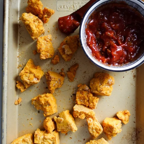 Chunks of crispy tofu nuggets on a baking tray, accompanied by a bowl filled with a red, chunky dipping sauce. An oven-fresh aroma wafts as a spoon rests in the bowl with its striped rim. Crumbs are scattered around, catching the light from a bright source.
